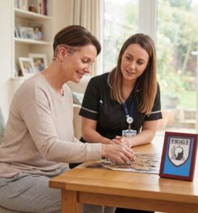 A smiling woman in casual clothes is seated at a wooden table in a bright living room, focusing on a jigsaw puzzle. Her hair is partially shaved on one side, revealing a visible, healing surgical scar behind her ear. Opposite her sits a healthcare professional in a dark uniform with an ID badge, observing and supporting the recovery activity. On the table is a framed shield logo featuring a brain, a spine, and the letters "RFG".